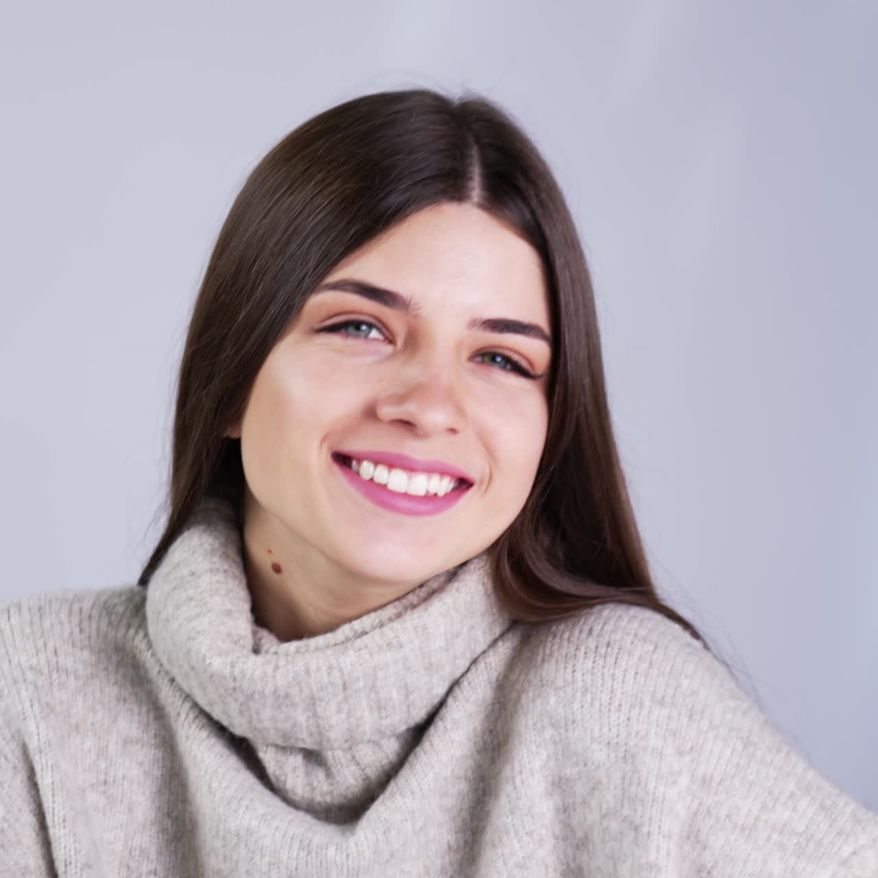 Cheerful smiling beautifully and touching her long dark hair. Female model wearing warm grey sweater posing in studio. Close up