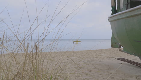 Leerer-Redlowo-Strand-Mit-Bewölktem-Himmel-In-Gdynia-Im-Winter---Landschaft-Einer-Ostsee-Durch-Trockenes-Gras,-Das-Neben-Einem-Alten-Rostigen-Fischerboot-Auf-Weißem-Sand-Steht