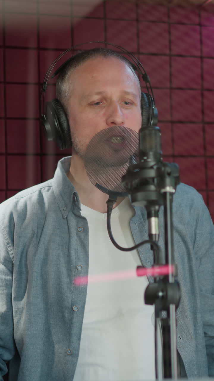 un hombre con una camisa de manga larga azul, camiseta blanca y auriculares negros está cantando apasionadamente en un estudio de grabación. está de pie frente a un micrófono con un panel acústico rojo