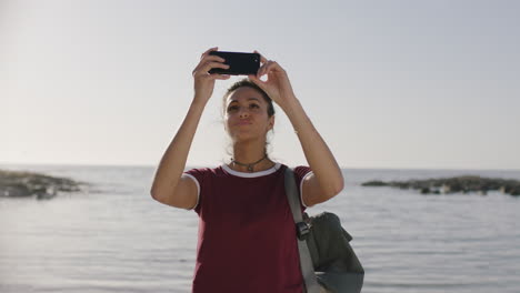 Retrato-De-Una-Joven-Hispana-Tomando-Fotos-En-La-Playa-Usando-Un-Teléfono-Inteligente-Disfrutando-De-Un-Día-Soleado-Y-Relajado