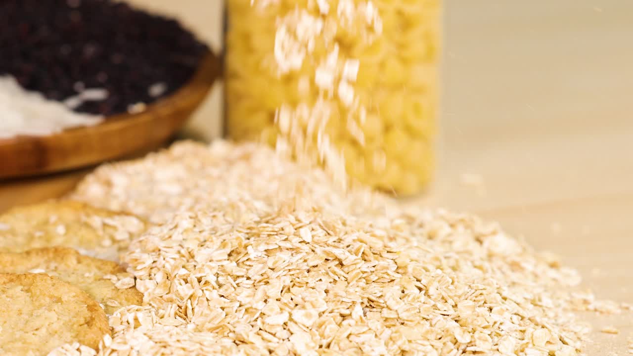 Rolled oats are poured from a glass onto a wooden surface, surrounded by jars of grains and seeds