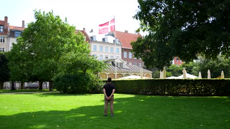 Back-view-of-man-looking-at-Danish-flag-waving-with-the-wind-in-Copenhagen,-Denmark