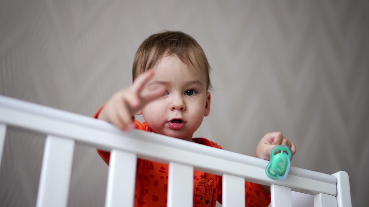 Cute one-year-old child standing in the crib and holding a pacifier in his hand. Baby looks straight into camera showing gestures with his hand.