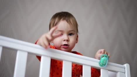 cute one-year-old child standing in the crib and holding a pacifier in his hand. baby looks straight into camera showing gestures with his hand.
