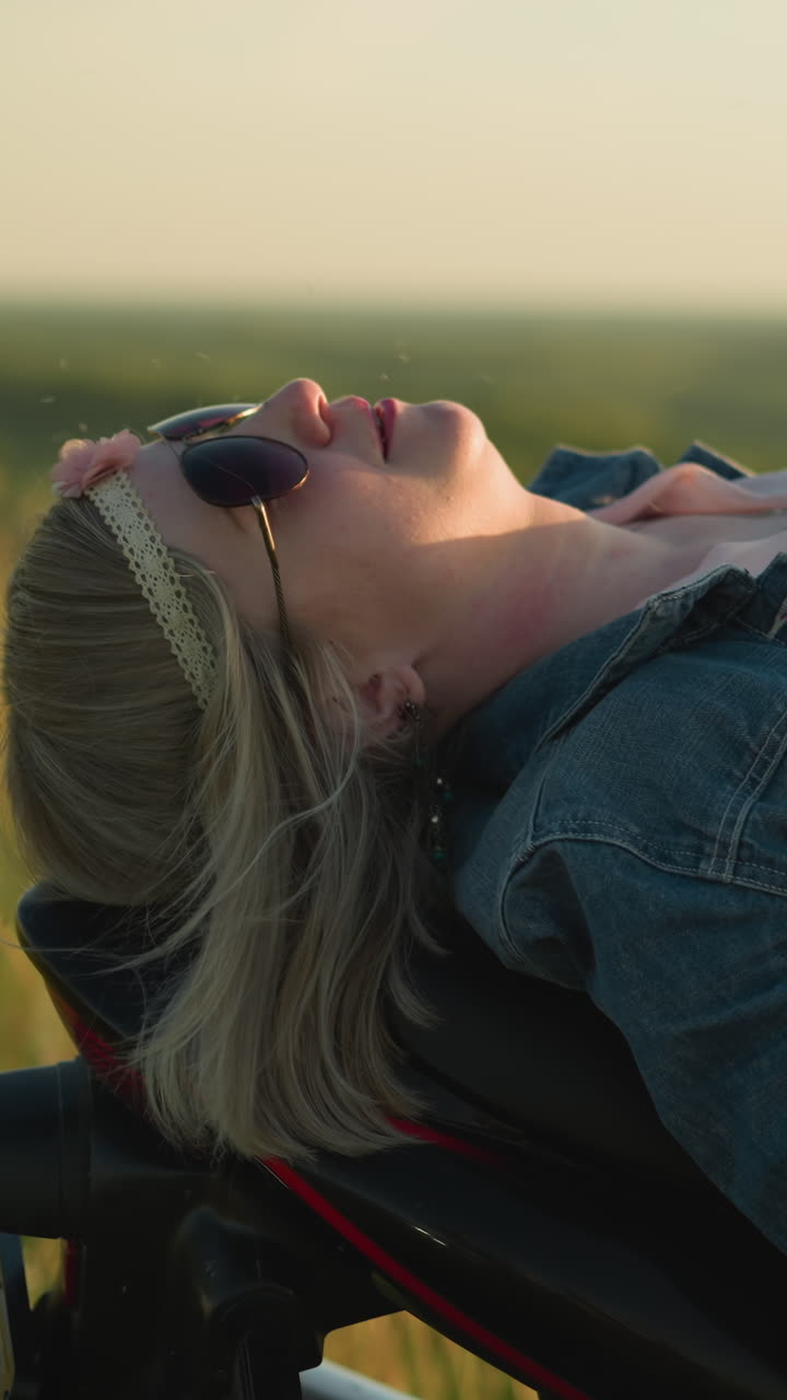 A woman lies back on her bike wearing sunglasses, adjusting her denim jacket as she relaxes in an open grassy field under the sun, enjoying the peaceful nature around her