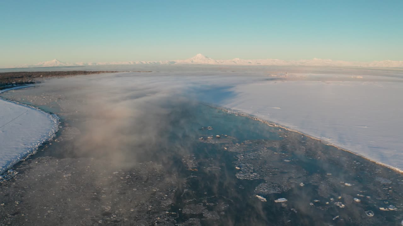Winter wonderland with a snowing cold river, clear skies, and misty fog rolling over the Kenai River in Alaska with Mountains in the background
