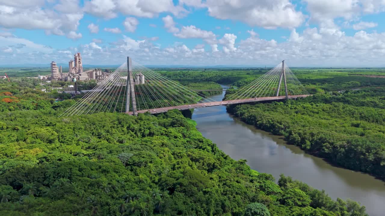 Mauricio Baez Bridge over river, with cement plant in background, infrastructure and industry surrounded by lush forest, Dominican Republic. Aerial forward