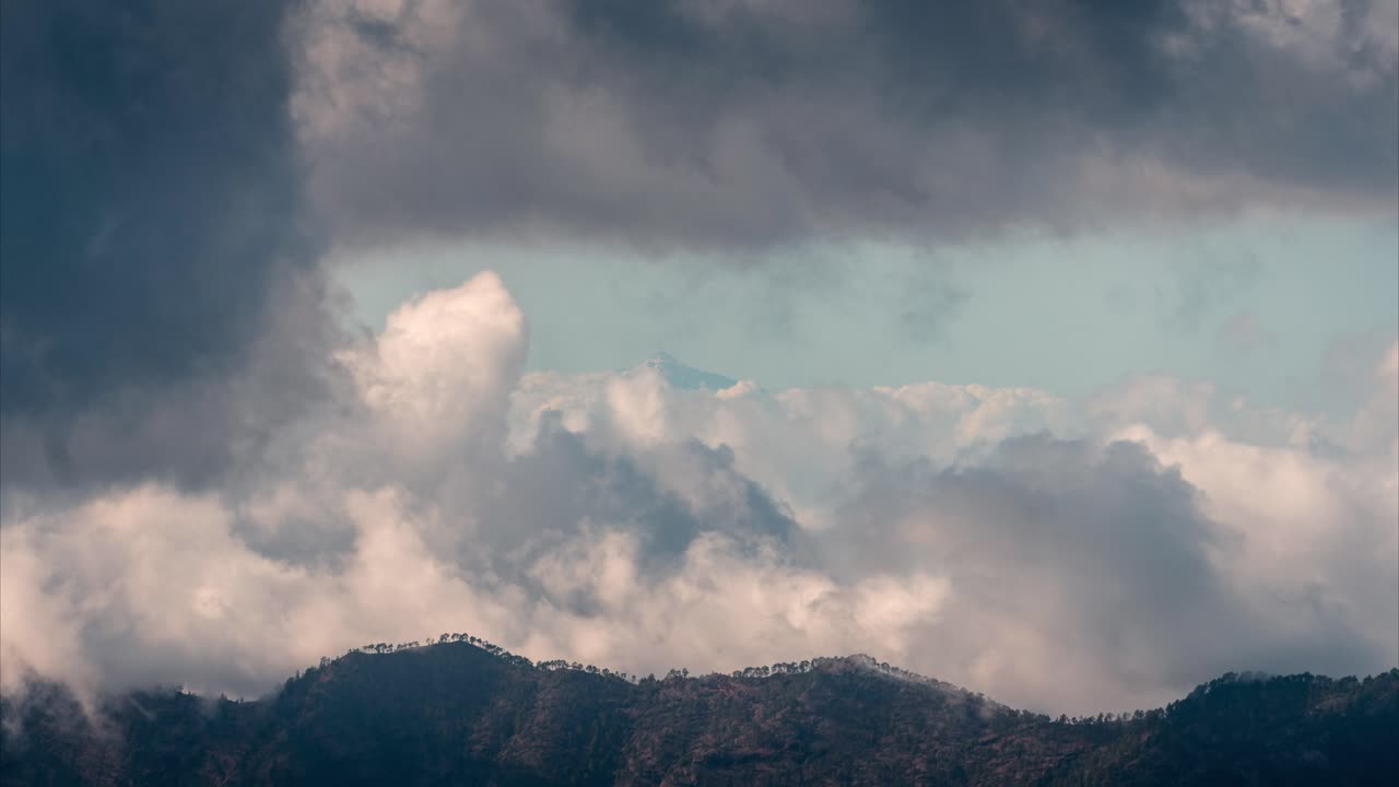 Distant view of Mount Teide covered in snow, rising above the clouds, seen from Gran Canaria on a moody winter day. A breathtaking contrast of volcanic peaks and dramatic sky.