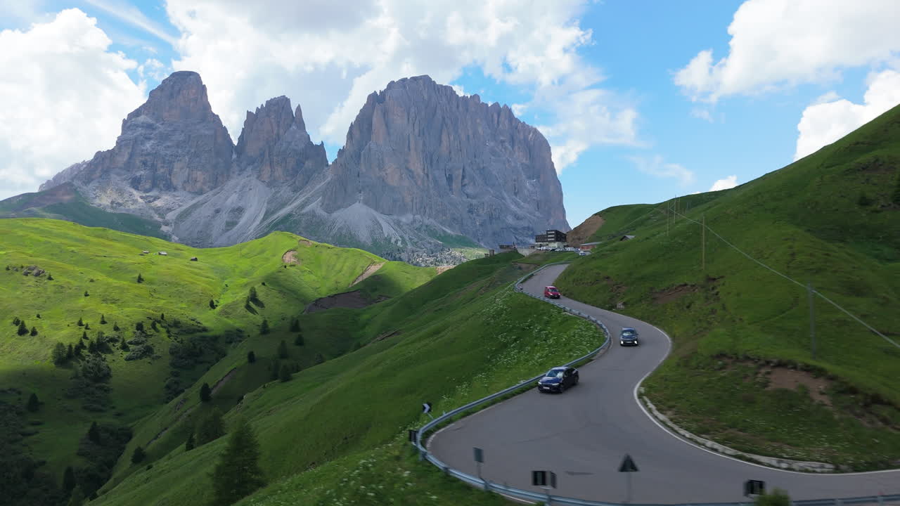 Aerial drone view of serpentine mountain road in the Dolomites with Grohmannspitze peak in background and moving cloud shadows across green slopes