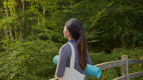 woman wearing sports clothing carrying yoga mat standing on bridge in forest looking around enjoying peace and beauty of nature