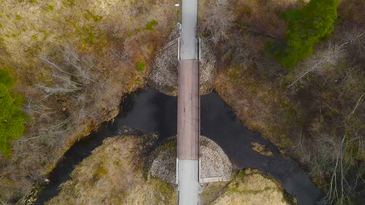 Top down aerial view decending upon a large wooden bridge that is going over a dark colored brown Laagri Pääskpla river during spring or autumn time cloudy day, leafless trees and brown nature around.