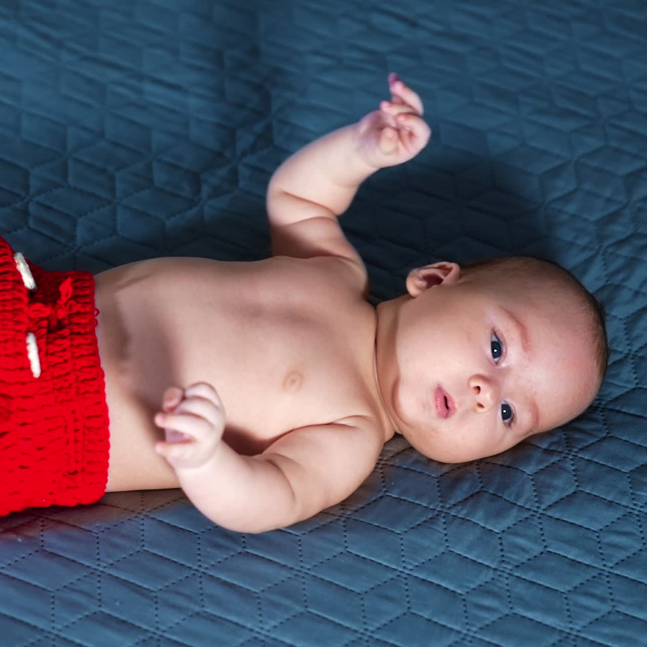 Active toddler boy in red shorts and white socks. Caucasian baby moves feet cheerfully while lying on the bed. Grey backdrop