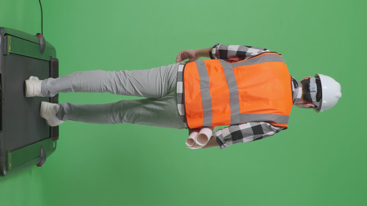 vista trasera de cuerpo completo ingeniero masculino asiático con casco de seguridad sosteniendo el plano en sus manos y mirando a su alrededor mientras camina en el estudio de fondo de pantalla verde