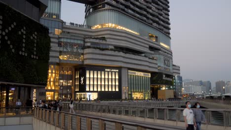 people walking on a promenade next to a modern shopping complex with luxury brand stores at dusk