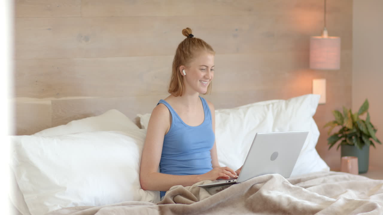 Using laptop and wearing earbuds, woman sitting on bed smiling, copy space