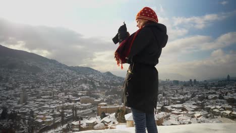 Mujer-Rubia-Parada-En-Una-Colina-Y-Tomando-Un-Video-De-Sarajevo-Con-Su-Teléfono-Móvil