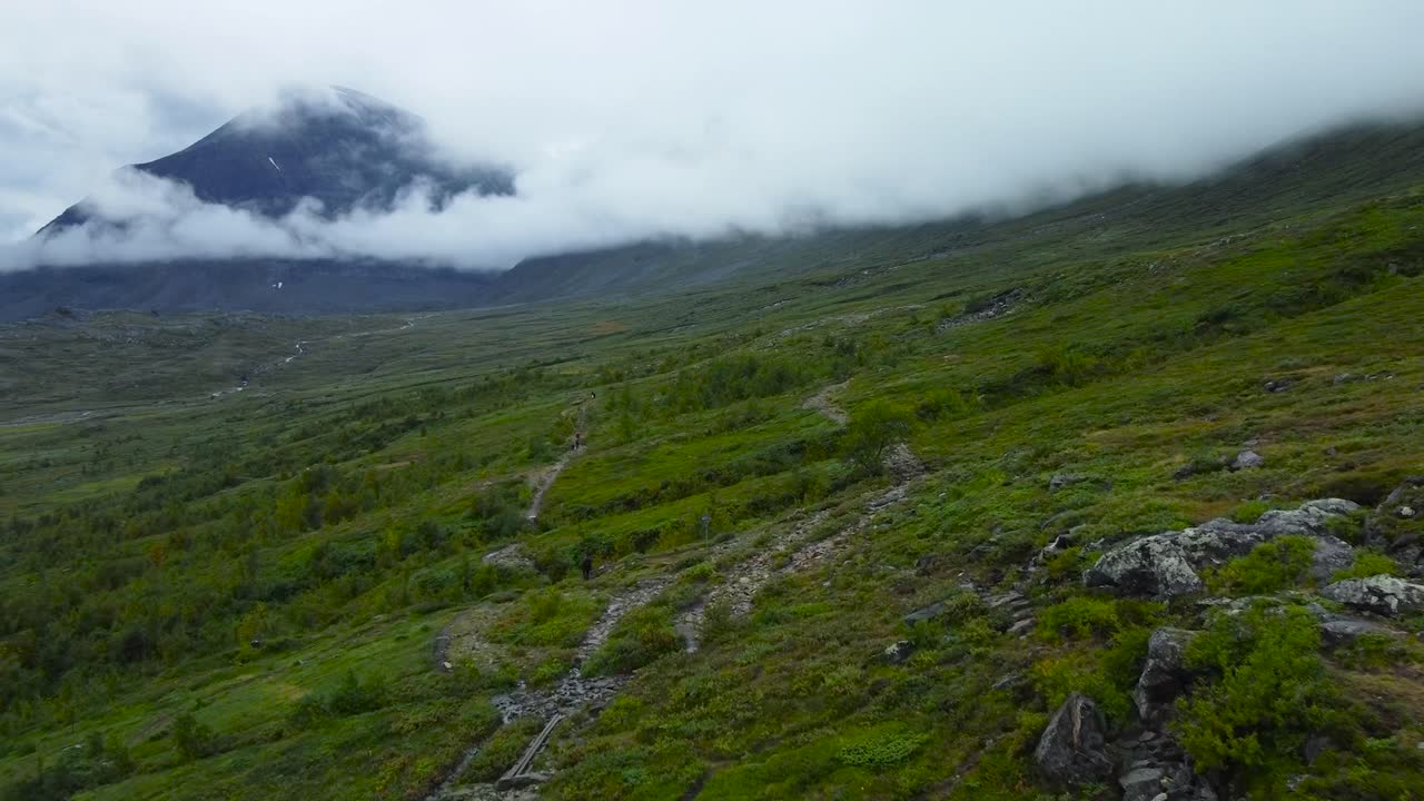 Aerial drone footage flying over green grassy and moss covered rocky slope terrain in sweden while a large black colored Kebnekaise mountain is visible submerged in white fluffy clouds in background.