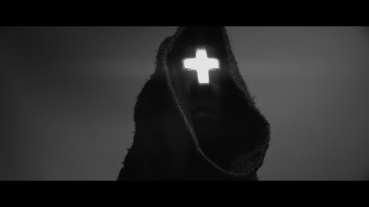 Masked and hooded man with cross of light glowing on his face looks in the camera