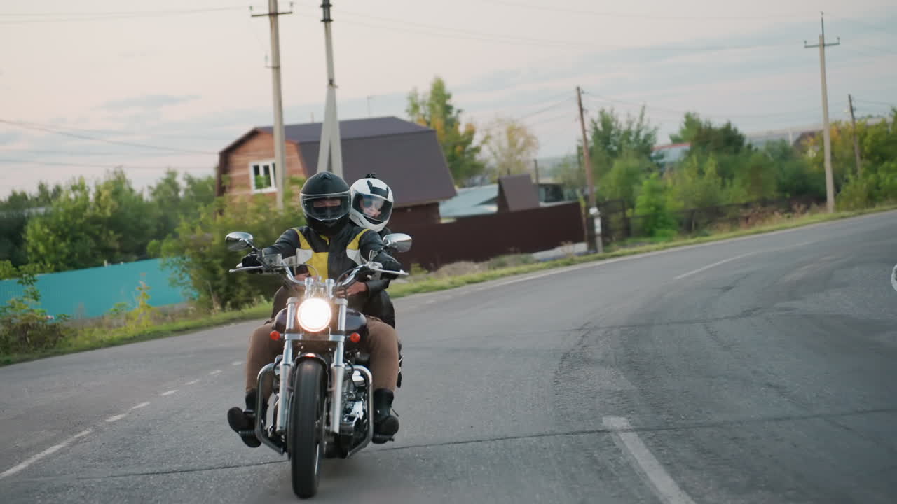 Motorcycle racer with passenger leans into bend on countryside road during trip, wearing helmets and protective gear, passing trees, utility poles, and rural buildings