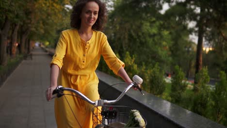 beautiful woman in long yellow dress riding a city bicycle with a basket and flowers in the city park looking in the camera and