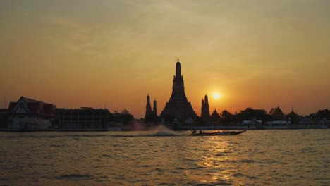 Wide-shot-of-Wat-Arun-at-Sunset-and-a-river-longtail-boat-drives-by,-Bangkok-Thailand