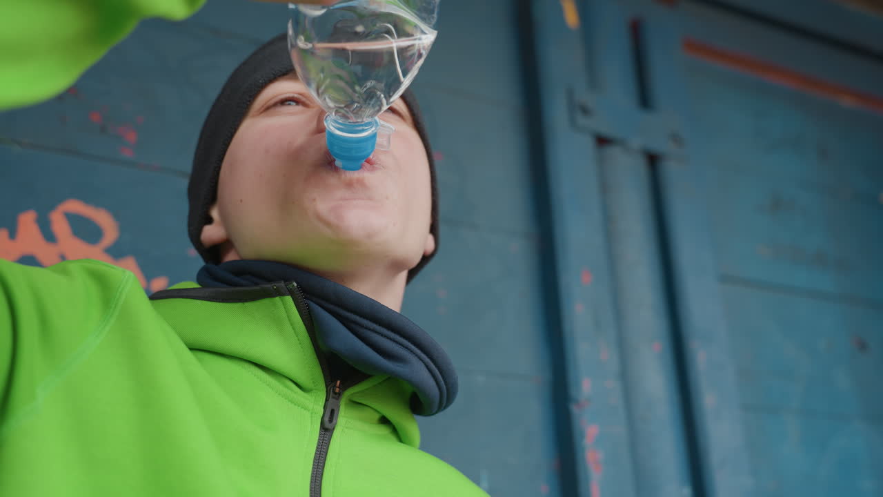 primer plano de un hombre caucásico con chaqueta verde bebiendo agua de una botella, escena invernal al aire libre con gorro de lana y bufanda, recuperación post-carrera y aliento visible, telón de fondo urbano con puerta azul y grafiti, enfoque selectivo