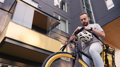 Food-Delivery-Guy-Wearing-Thermal-Backpack-Recorded-From-Below-Puts-On-A-Helmet-And-Start-Riding-His-Bike