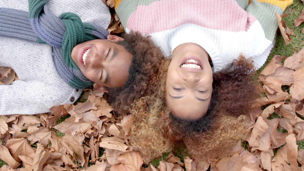 Happy african american mother and son in warm clothes lying on fallen leaves in garden, slow motion
