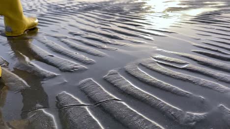 Sun-reflection-on-a-rippled-sandy-beach-and-yellow-boots-walking-in-slow-motion