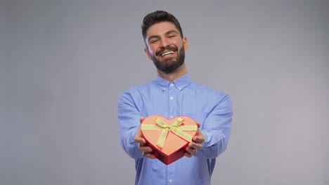 Joven-Feliz-Con-Caja-De-Regalo-En-Forma-De-Corazón.-Día-De-San-Valentín,-Saludo-Y-Concepto-De-Vacaciones:-Retrato-En-Video-De-Un-Joven-Feliz-Y-Sonriente-Con-Una-Caja-De-Regalo-En-Forma-De-Corazón-Sobre-Fondo-Gris