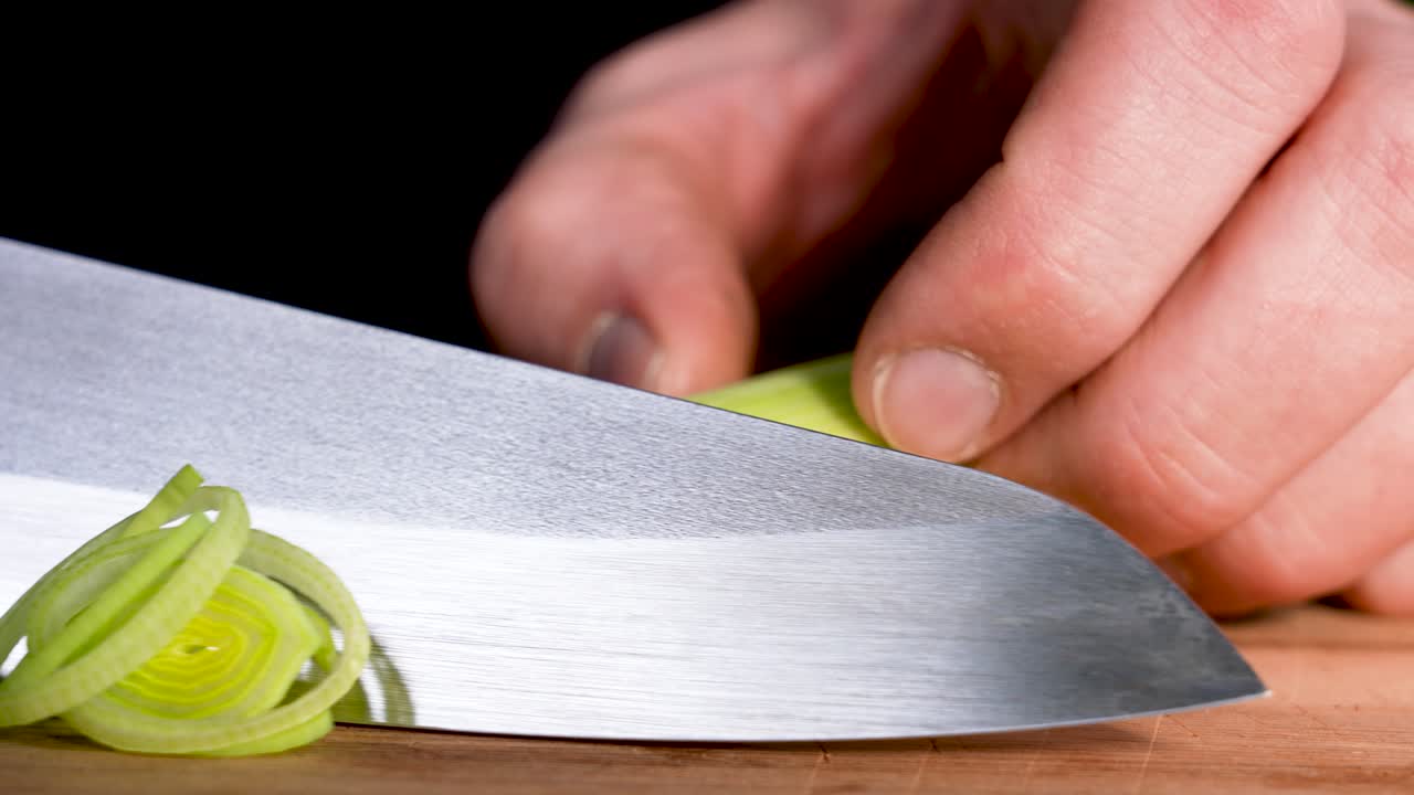 A sharp kitchen knife cuts cleanly through a fresh leek in a detailed macro close-up. The crisp green layers and smooth motion highlight precise food preparation and natural culinary visuals
