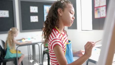 biracial girl with brown hair focuses on painting in a classroom in school