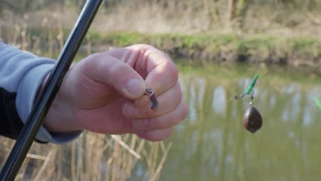 hand holding baited hook in nature, fishing rod in river background