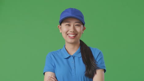 푸른 유니폼을 입은 아시아 여성 택배사가 스튜디오의 녹색 스크린 배경에서 팔을 는 웃음을 짓고 있습니다.