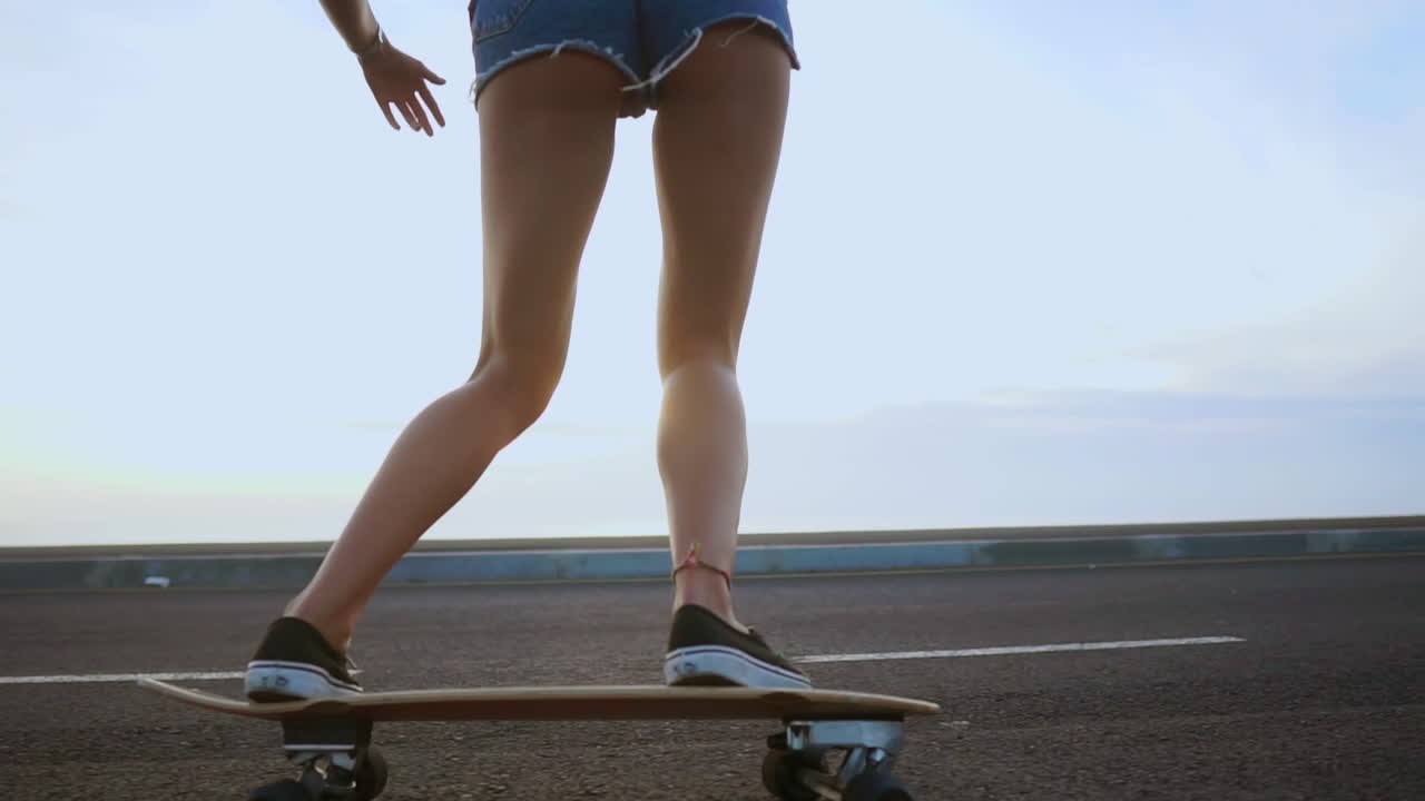 A slow-motion scene captures a woman skateboarding on a road at sunset, framed by mountains and a beautiful sky. She's wearing shorts