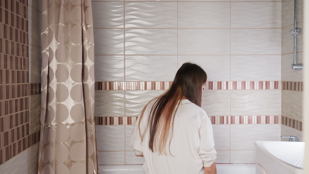 Back view of fair skinned girl enters bathroom, walks to bathtub, lifts shower, sits on tub rim, washes feet near curtain, soft light on tiles, quiet hygiene routine, calm mood, gentle motion