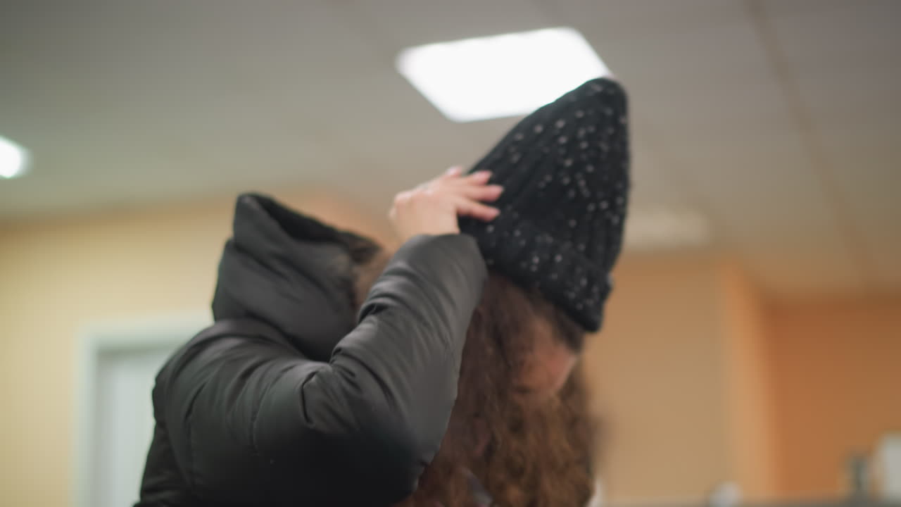 Girl in black winter jacket bends forward tossing long curly hair, hands lifted in motion, scarf draped over shoulders, candid indoor scene as she wears her cap natural beauty in expressive gesture