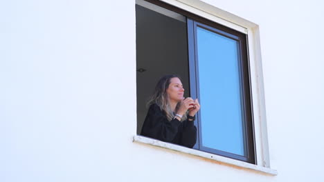 medium shot of young blond woman on window drinking coffee or tea and waving at neighbor