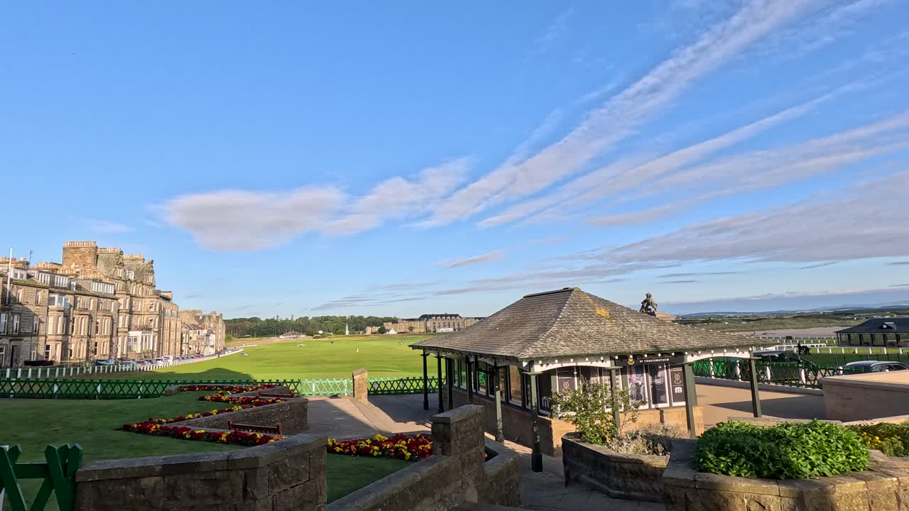 Wide pan reveals traditional golf clubhouse, manicured grounds, parked cars, and clear summer sky