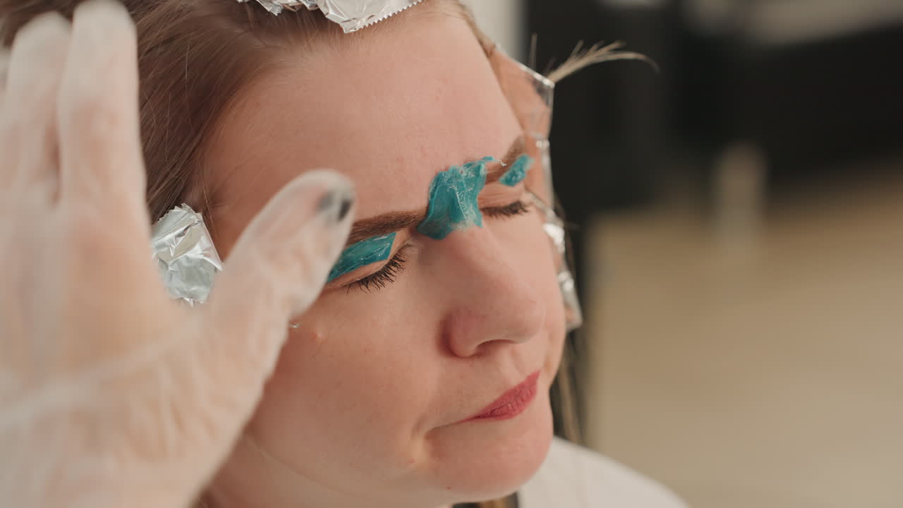 salon eyebrow tinting blue pigment application esthetician wearing gloves applies turquoise pigment across brows while foil protects hairline, client relaxed in chair, close facial detail, precise