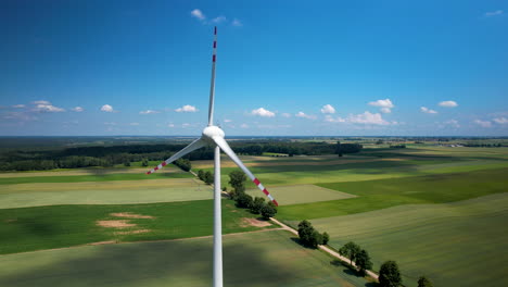 Aerogenerador-Girando-Lentamente-En-El-Campo-Con-Tierras-De-Cultivo-Verde,-Antena