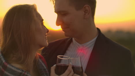 A-young-couple-on-the-roof-drinking-wine-from-glasses-standing-in-a-blanket-and-admiring-the-beautiful-sunset-over-the-city.-Romantic-evening-on-the-roof-overlooking-the-city