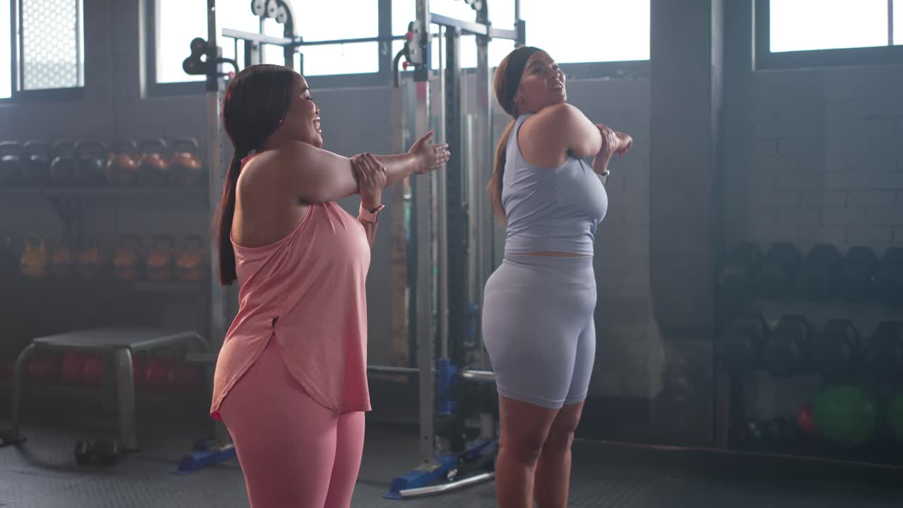 Diverse female friends following leader's crossbody shoulder stretch by gym weights, warming up