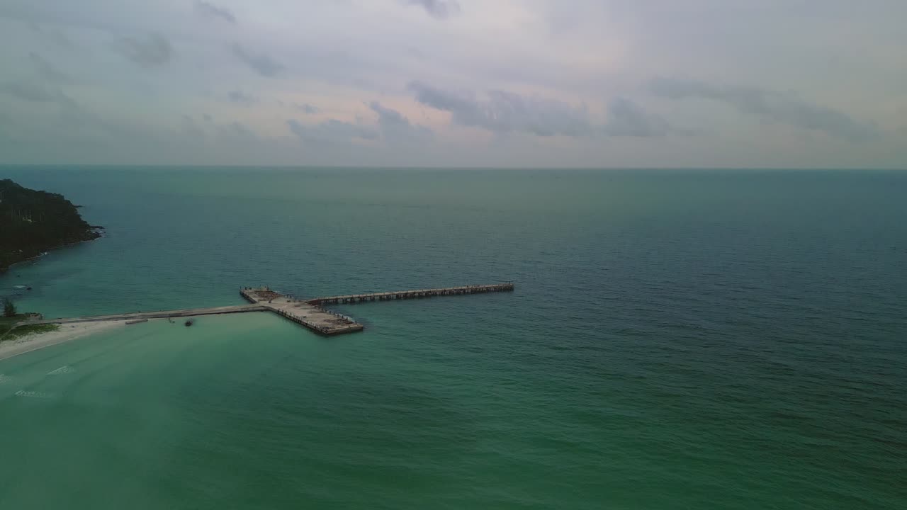 A concrete pier stretches out from the beach into the dark ocean under a cloudy sky.