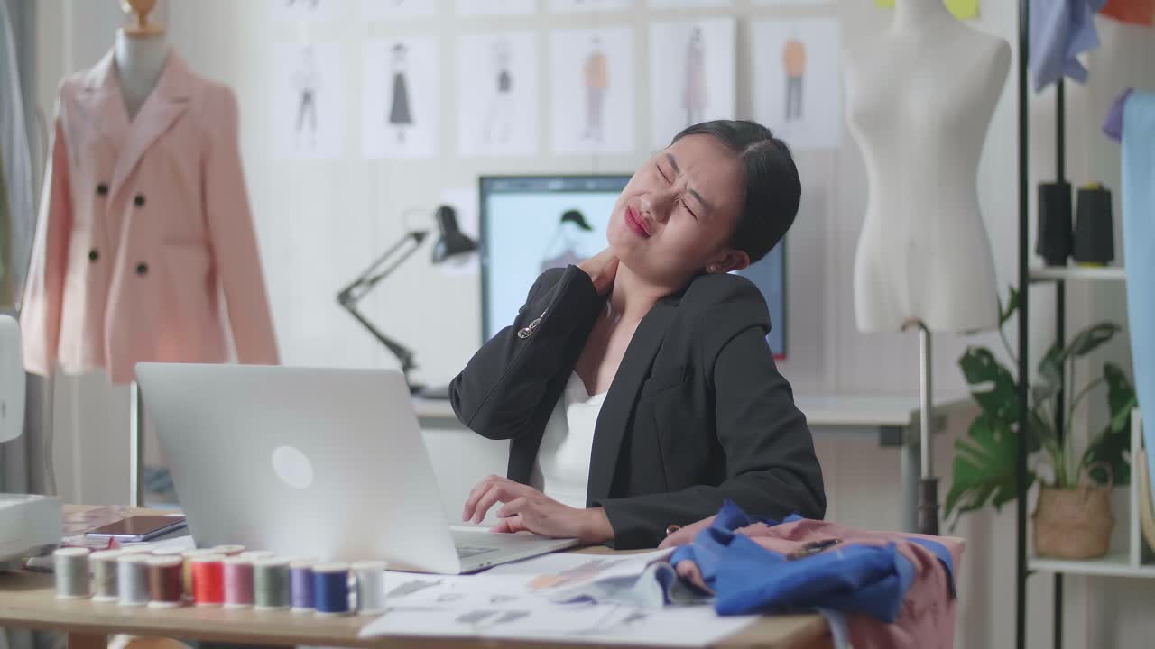 비즈니스 슈트 를 입은 아시아 여성 디자이너 가 노트북 을 사용 하고 스튜디오 에서 옷 을 디자인 하는 동안 스트레칭 을 하고 있다