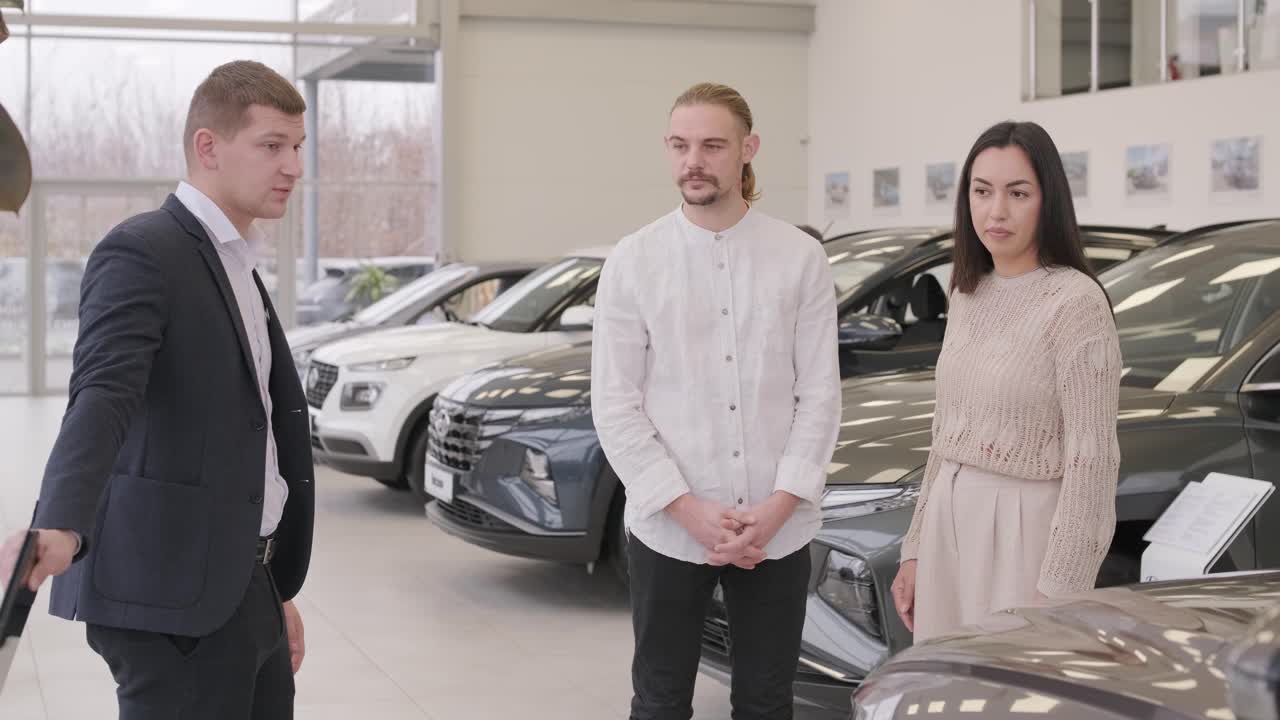Young car salesman showing to young couple new automobile at dealership salon.