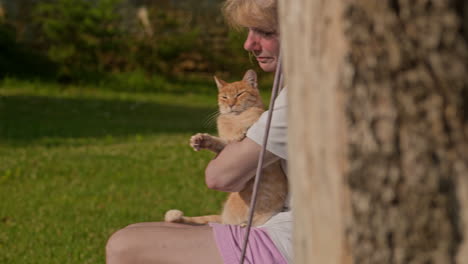 la mujer en el columpio del árbol acaricia a su adorable gato pelirrojo, revela la diapositiva