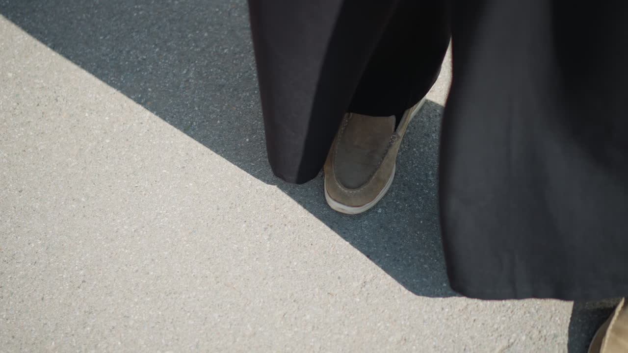 Top close up view of lady legs shifting slightly on bright sunlit pavement, wearing black pants and brown shoes, subtle movement and shadow convey worry, hesitation, and quiet emotional tension