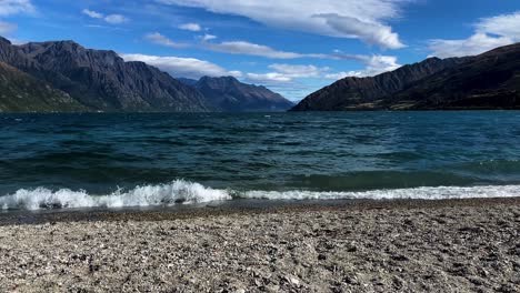 Calm-waves-crashing-against-rocky-lake-shoreline,-with-mountains-and-blue-sky