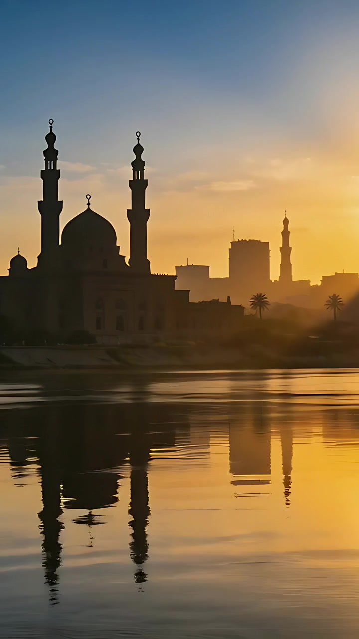 Video verticale: Sole nascente che illumina la cupola di una moschea e due minareti sul fiume, riflettendosi sull'acqua calma.
